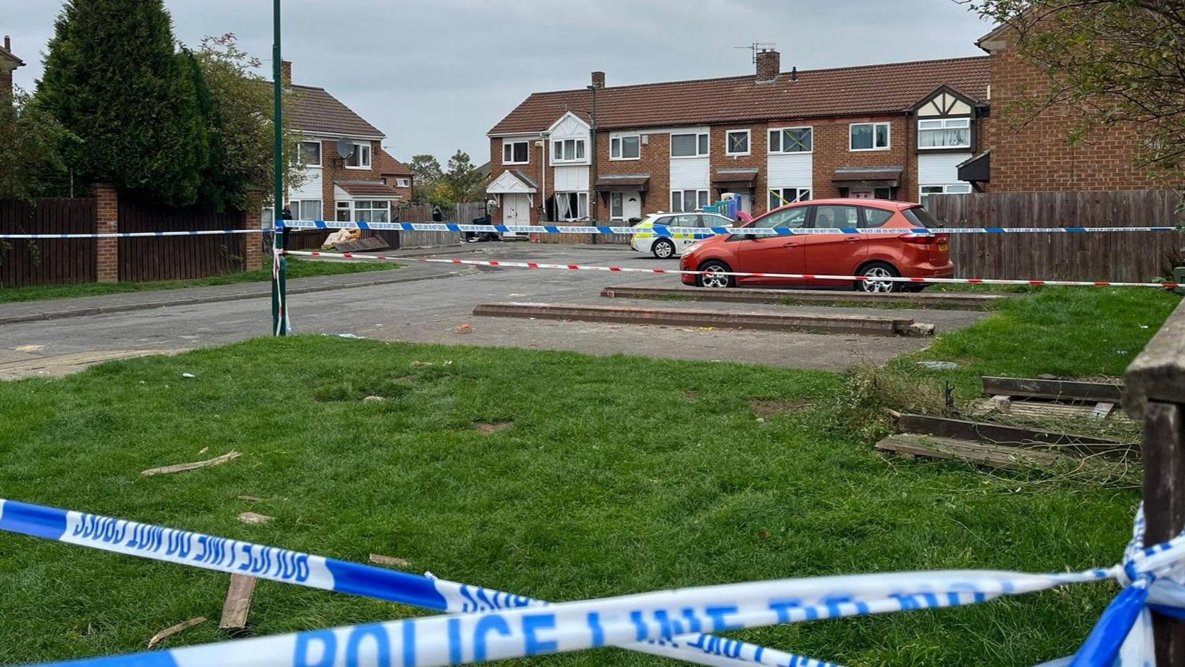 Richmond Court in Grangetown: a small cul-de-sac consisting of a cluster of red-brick houses. Police tape has been hung across the road to prevent access. A police car is parked outside a house which has a broken window.