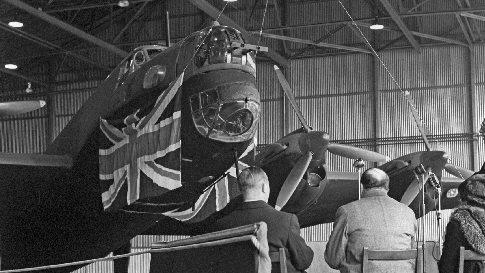 A black and white picture of a Halifax bomber in a hangar. There is a Union Jack flag hanging across the fuselage