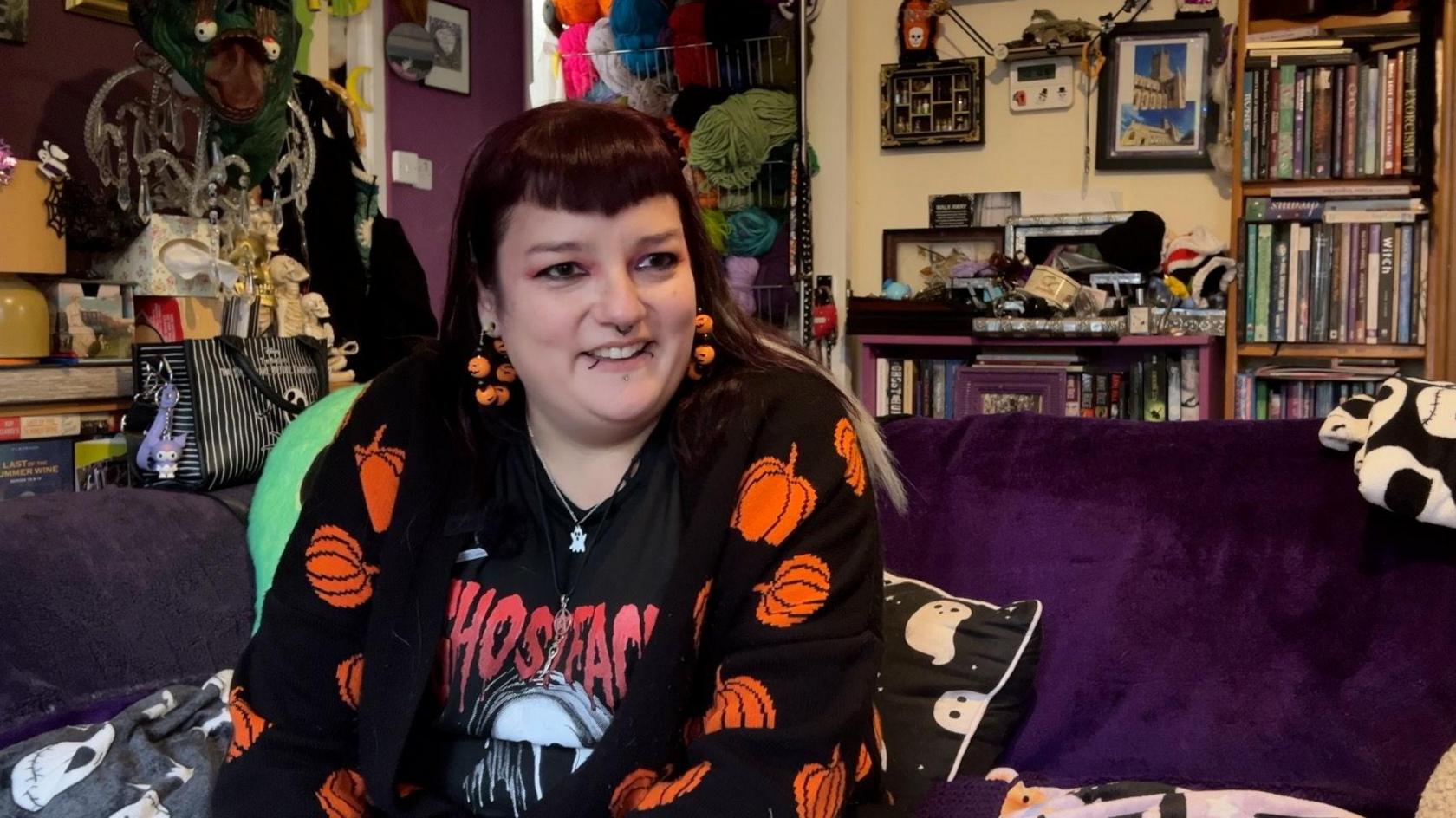 A young woman with long straight purple hair and a pointed full fringe smiles, looking out of frame. She wears a black cardigan with orange pumpkins on it, a Scream film t-shirt and pumpkin earrings. Her sofa has a dark purple blanket covering it and the living room behind her is full of decorations.
