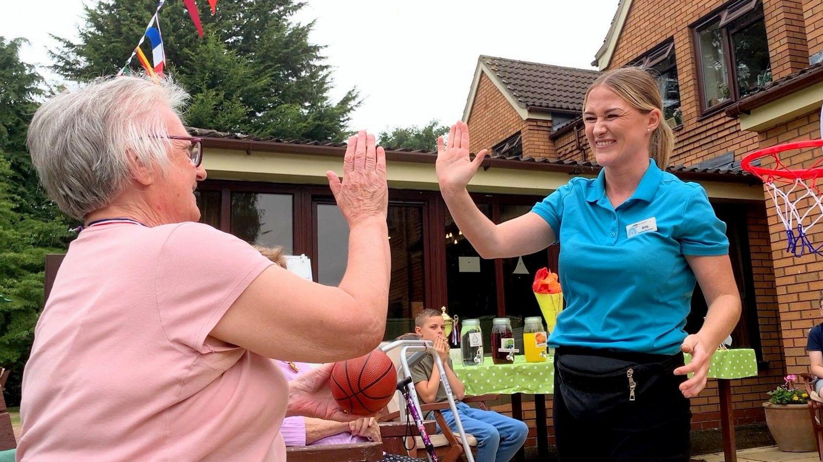 Amy Robinson has light brown hair and is wearing a turquoise top. She is high-fiving Pauline, who is wearing a pink top and glasses