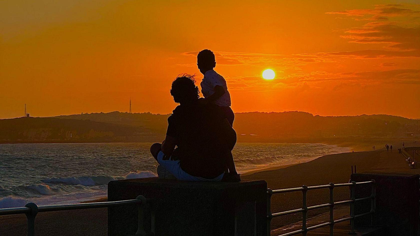 A silhouetted man holds his son on his shoulder in front of a sunset. 