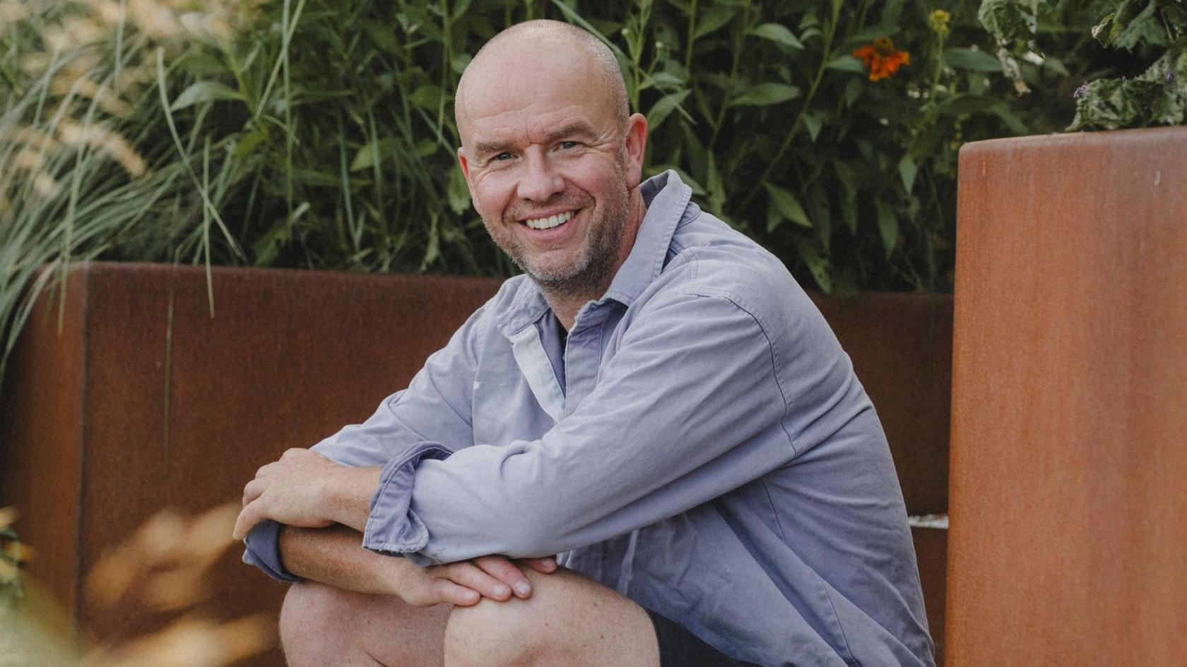 Darren Hawkes sits on a step in the midst of a garden with copper planters. He is wearing a blue shirt and has shorts on.