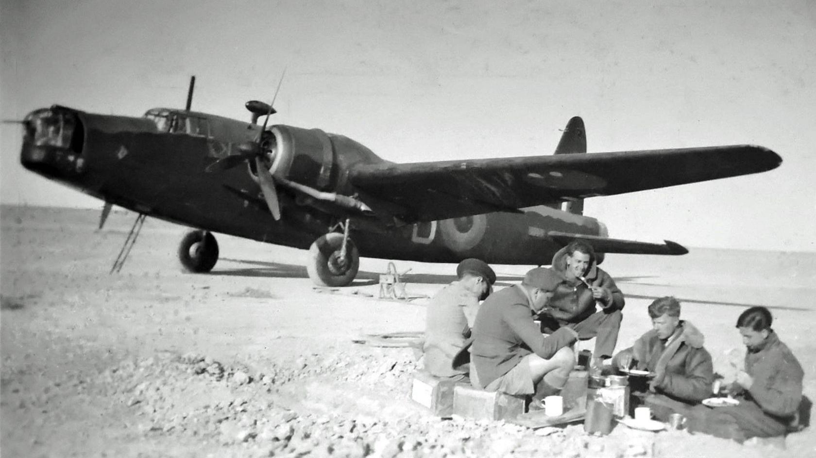 A black and white photograph showing a group of five men in uniform sitting on bar ground eating. Behind them is a Wellington bomber.