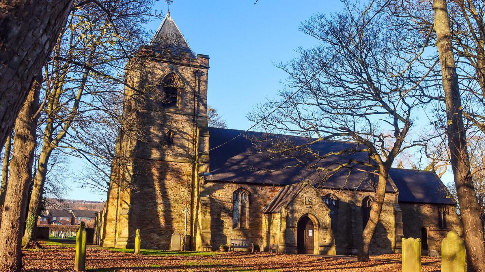 The frontage of St Stephen's Church in Willington on a sunny winter day. There are thousands of brown leaves in front of the church and the trees are bare. The church is a classically old building with gravestones outside in the yard. 