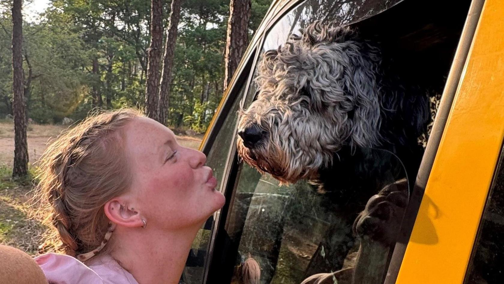 Ella Dungey has her fair hair in a plait. She is wearing a pink shirt and pouting at her dog, which is a black and grey bernedoodle and is poking its head out of a window on the yellow van. They are parked in a wooded area.