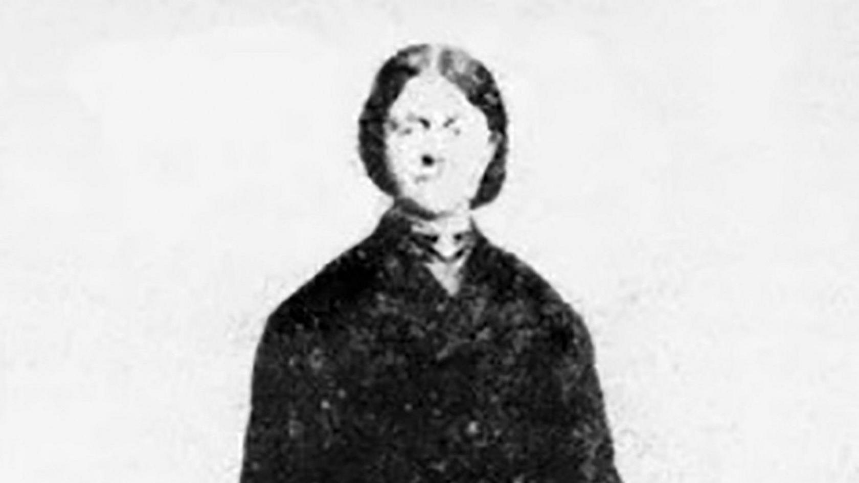 A primitive photograph of Emily outside her Plas Castell home. She is wearing a black coat and holding a hat. Her dark hair is swept back in a bun