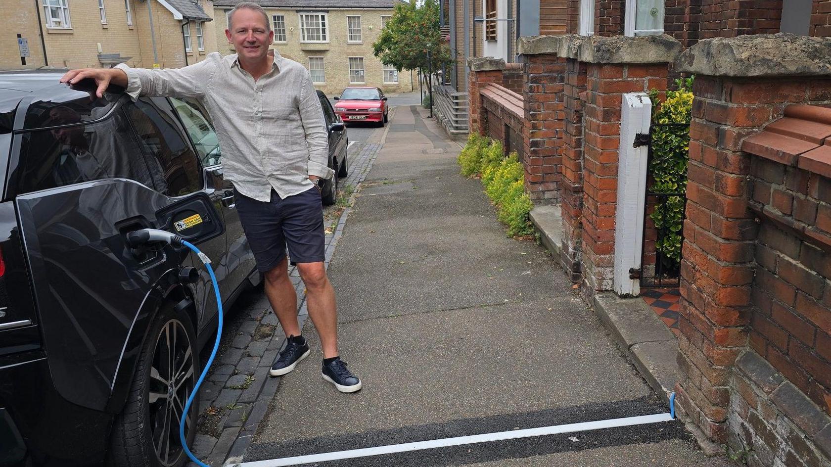 A man stands on the pavement leaning on his black electric car. He is wearing navy shorts and a beige long-sleeved shirt. The car is parked on the street, and a blue charging cable runs into a gully through the pavement, covered by a narrow metal strip. It connects to the wall of a terrace house which has red brick walls. Other houses and buildings are in the background, as are a few parked cars.