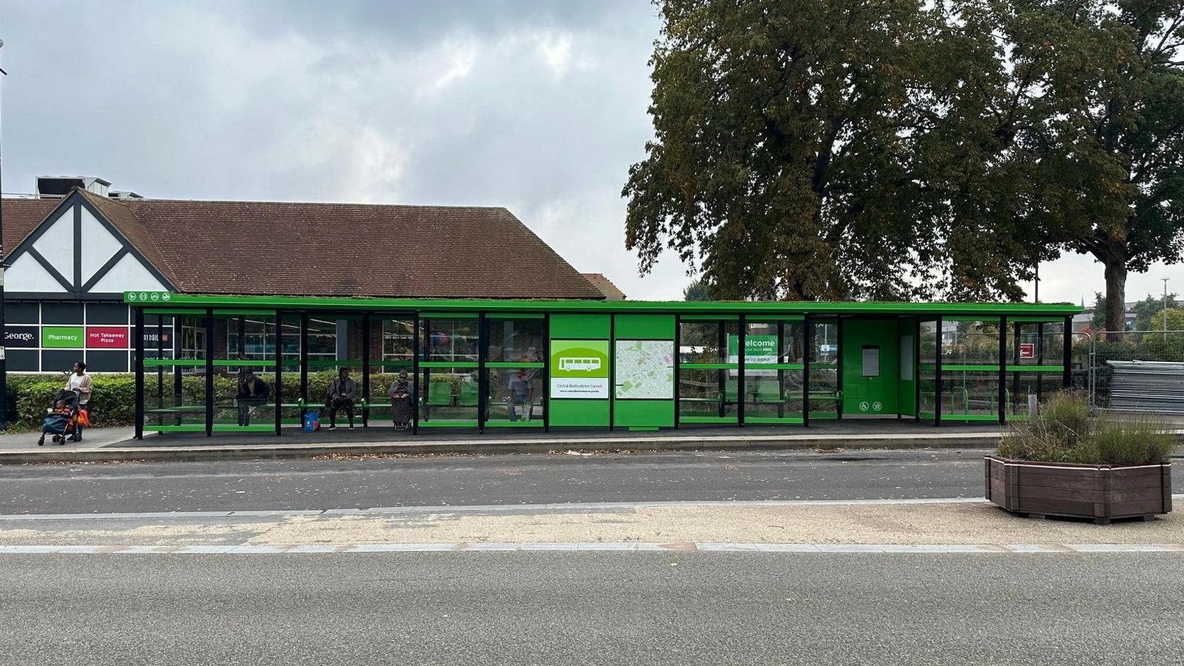 A bright green very wide bus shelter in front of a supermarket and large tree