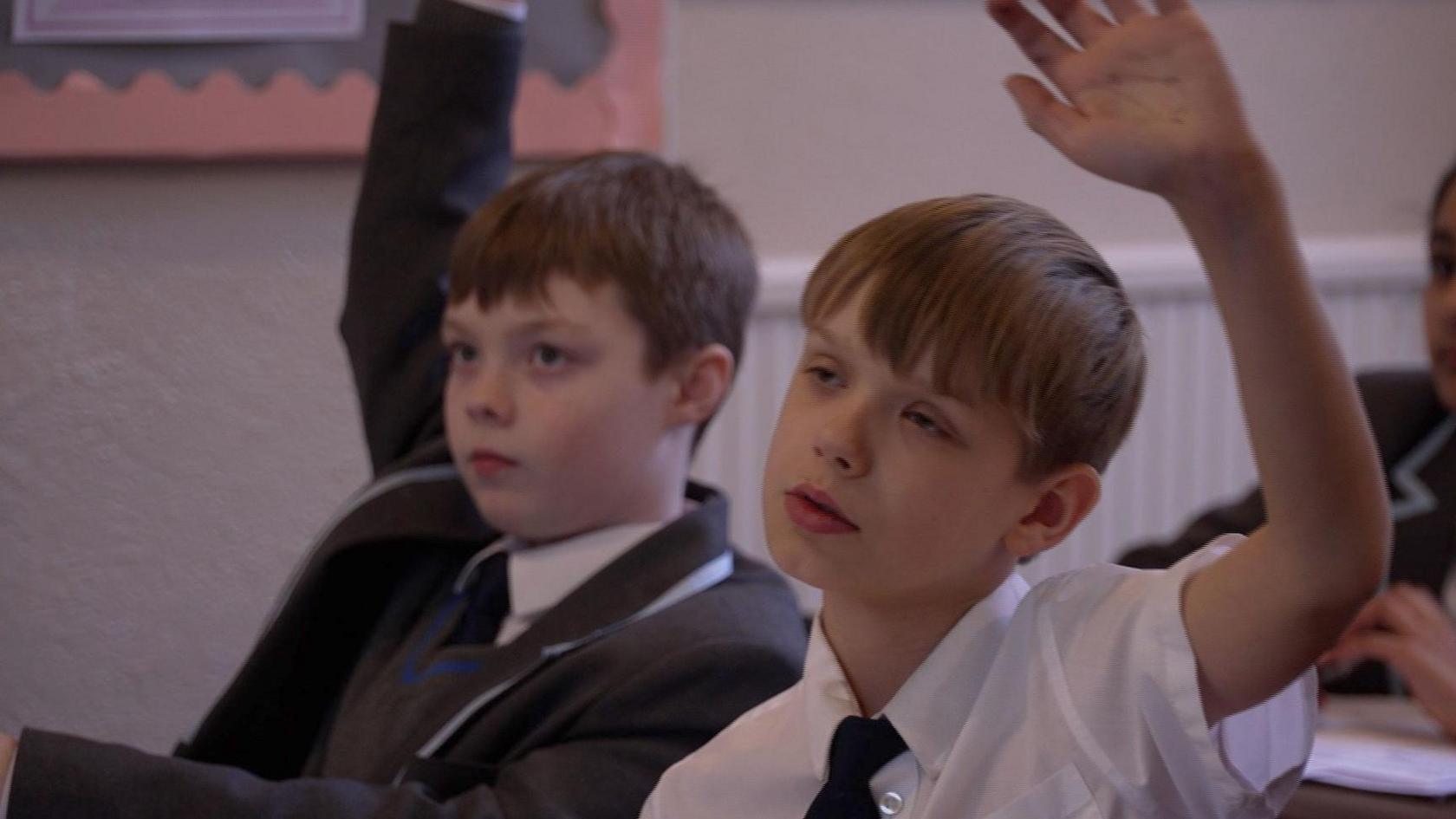 Two boys - aged around 10-11 are sat in a history class with their hands up. They are both wearing smart white shirts, with a tie and the boy on the left is wearing a deep grey blazer.