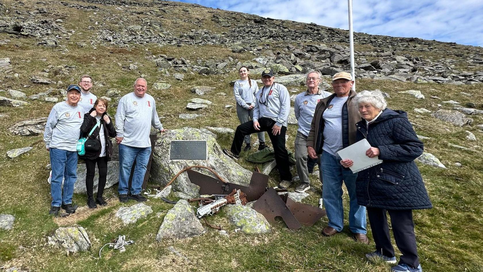 The group of family and friends and local guides at the North Barrule crash site memorial plaque, next to a flagpole.