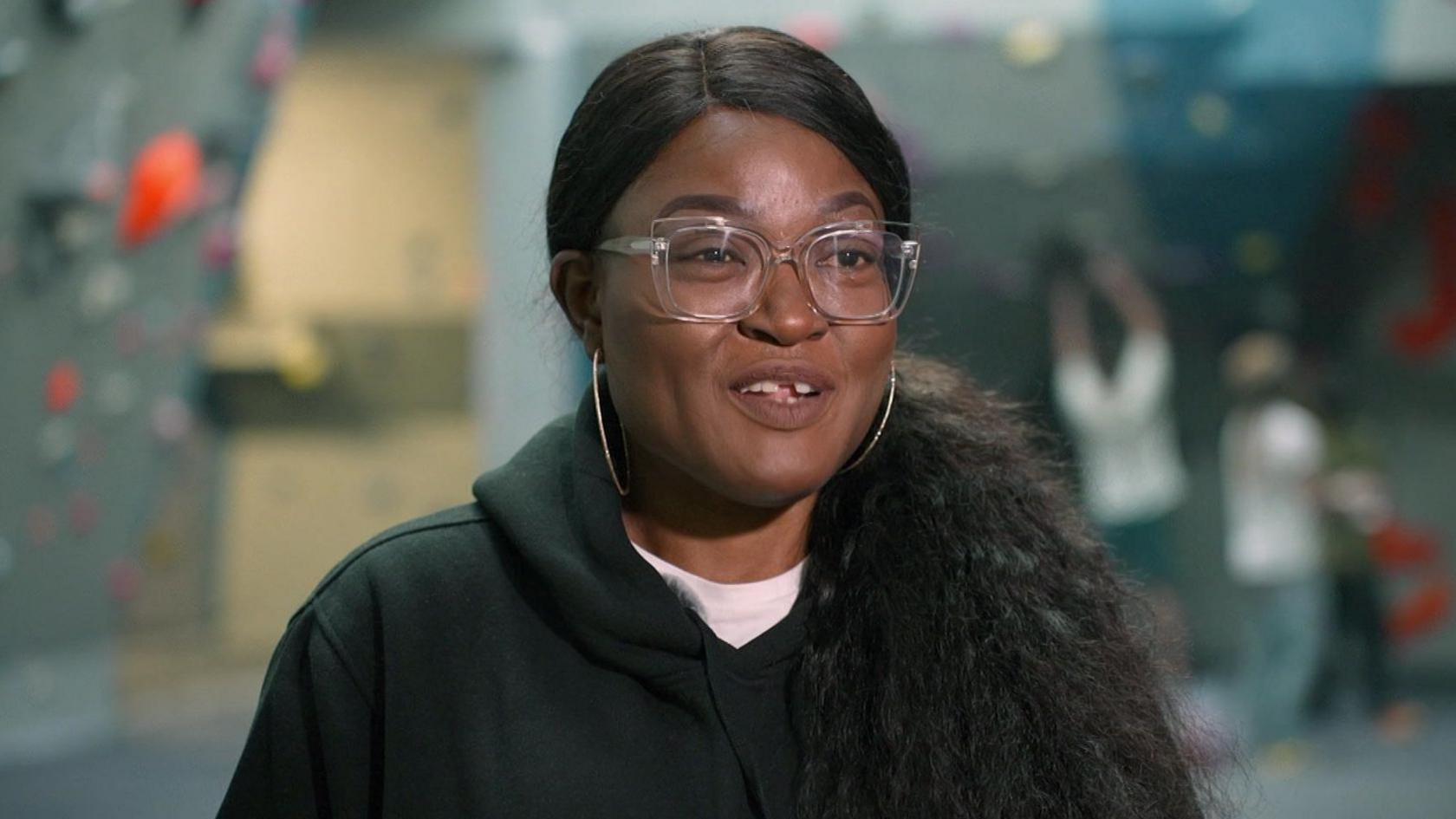 A woman is smiling as she talks to the camera while being interviewed. She is wearing a black top. She has long black hair which is tied back and is wearing glasses. A blurred out climbing wall with people climbing on it can be seen in the background.