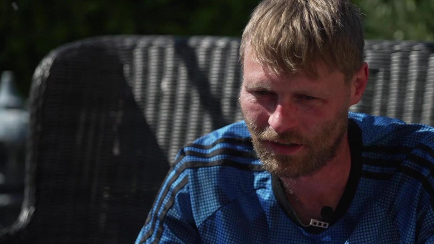 A man with blonde hair and a bear, wearing a blue football shirt, wears a serious expression as he sits on a wicker chair in a garden.