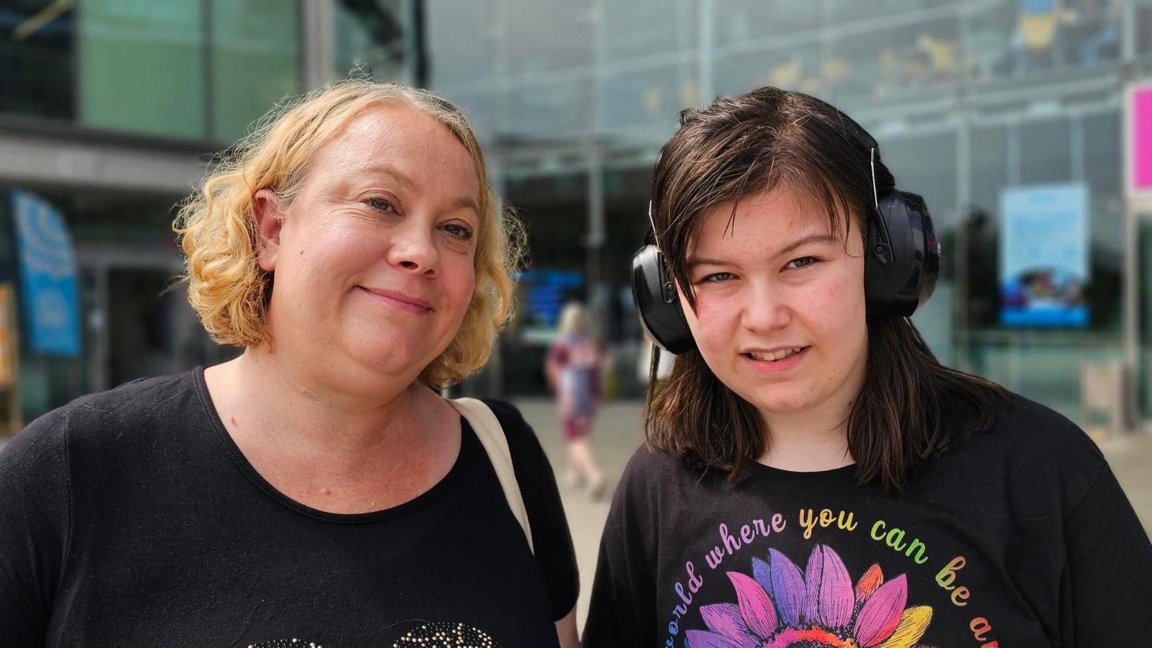 A closed mouth smiling Victoria Trattles who has blonde chin-length hair, wearing a black t-shirt with her daughter Maria who has dark shoulder-length hair and is wearing ear protectors and a black t-shite with a multi-coloured flower on it 