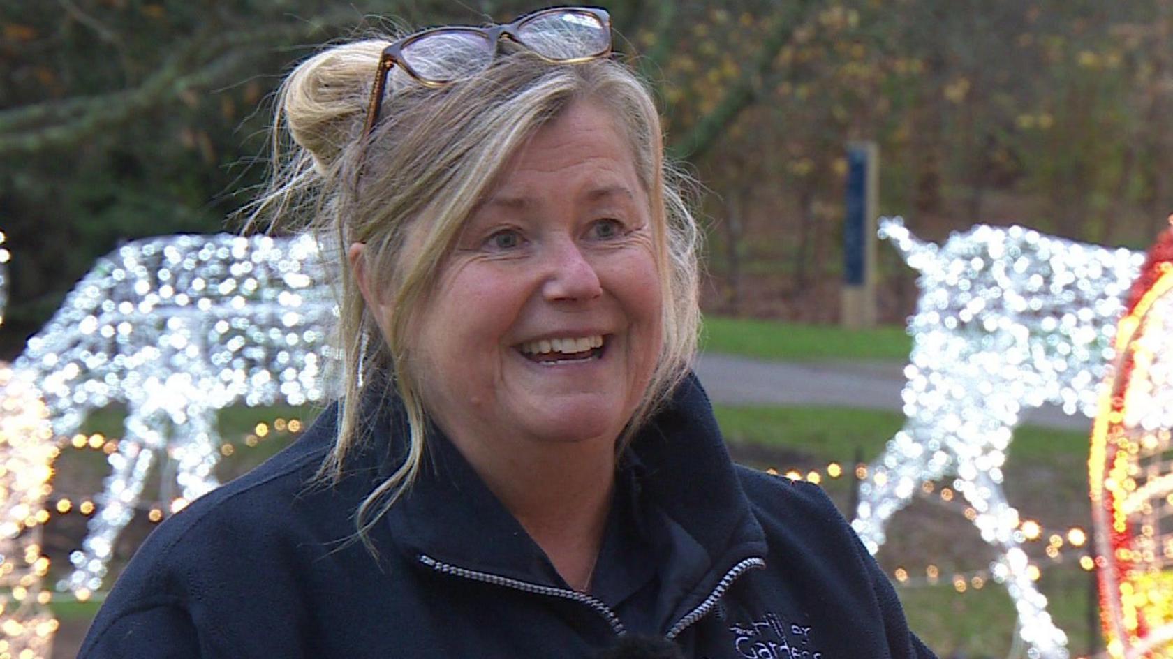 Tracy Randall looks slightly off camera as she gives her interview. She has blonde hair, which is tied back in a bun and a side fringe framing her face. She has a pair of thick framed tortoiseshell glasses on her head and wears a navy blue fleece with the Hillier's Garden logo on it. Behind her are fairy light sculptures of reindeer, which are slightly out of focus.