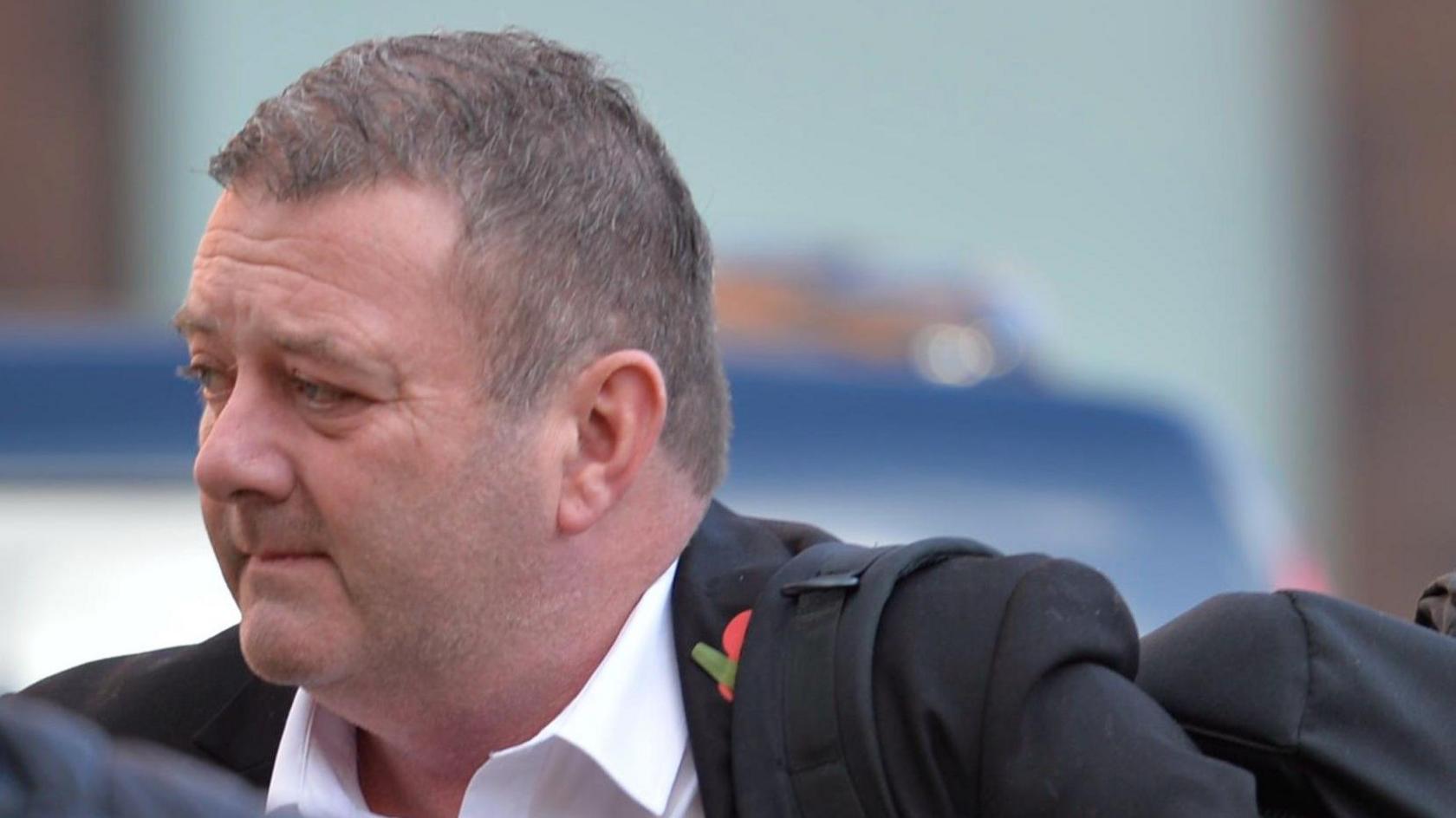 Alan Sharp looking away from the camera. He has short, grey hair. He is wearing a black suit over a white shirt and has a poppy under a black rucksack strap on his upper left chest.