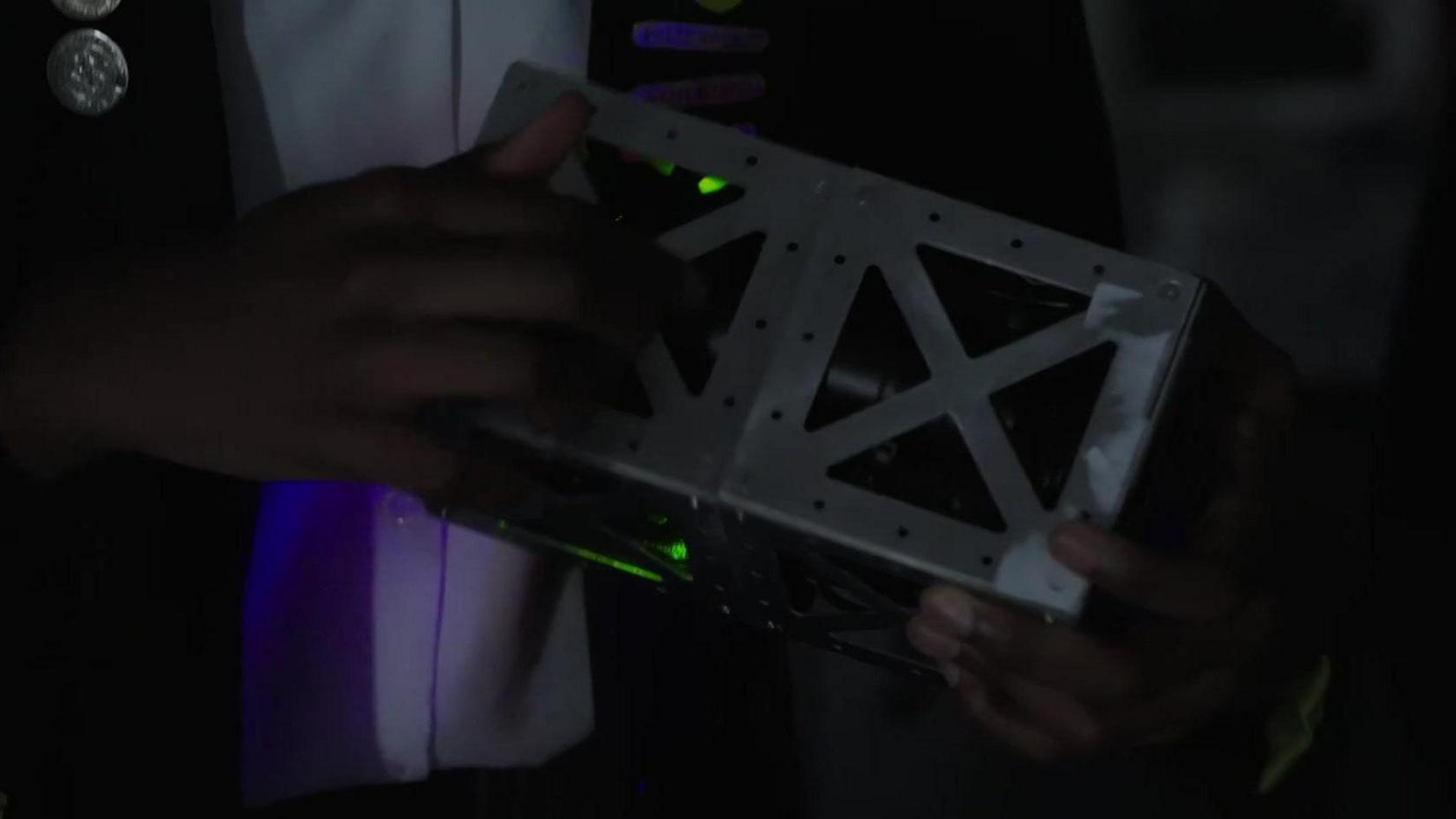 A close up of the satellite's metal casing - it is held in the hands of one of the pupils in a darkened room and it has some small green lights on the inside of the casing. 