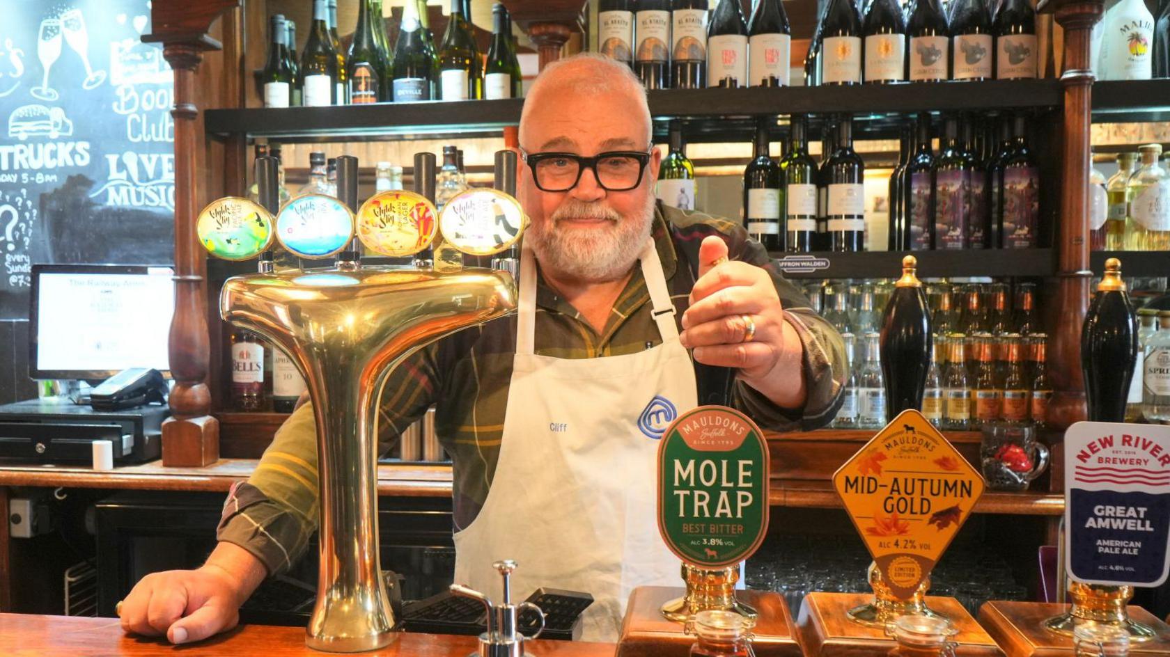 Cliff Parisi, wearing thick black glasses, pours a pint of beer behind while stood behind the bar