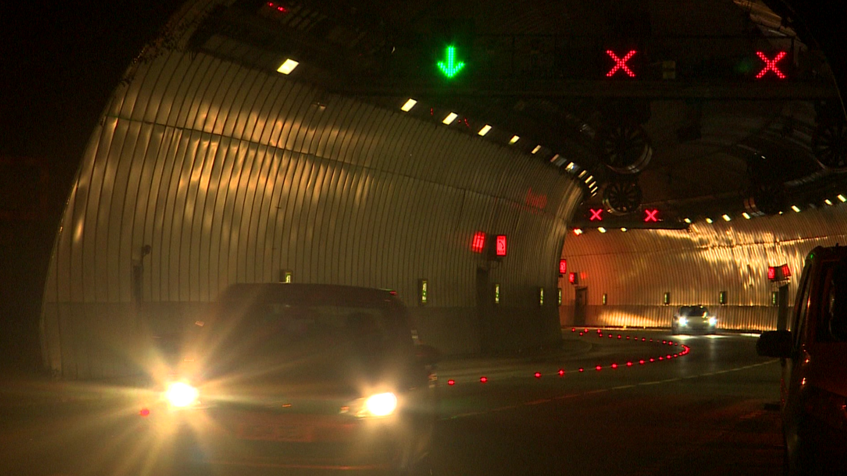 Several cars with headlights on drive through a dark, curved road tunnel with illuminated green arrows and red crosses above their heads.