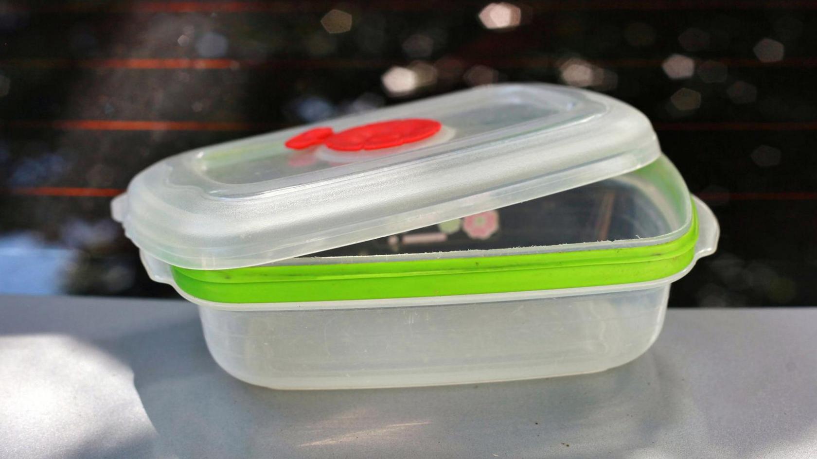 An empty plastic lunchbox. It is translucent with a lime green rim on the main box. The lid is slightly off and has a red vent on the top. It is placed on a worktop with a blurred background. 