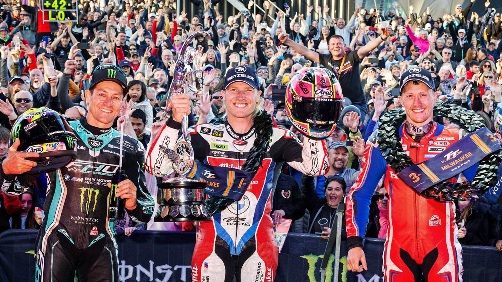 Three smiling men in motorbike leathers wearing garlands standing in front of a crowd of people, with the rider in the centre holding up a silver trophy