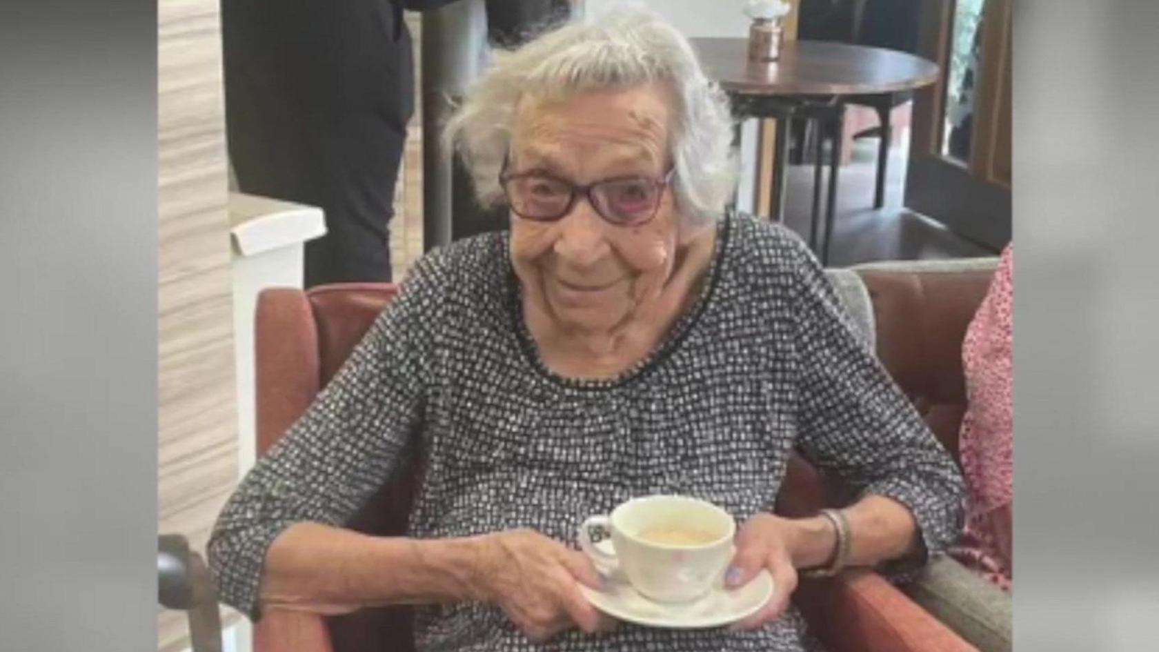 Betty sits in a red chair while holding a cup of tea and smiling as she looks at the camera. She is wearing a grey patterned top and there is a round table behind her.