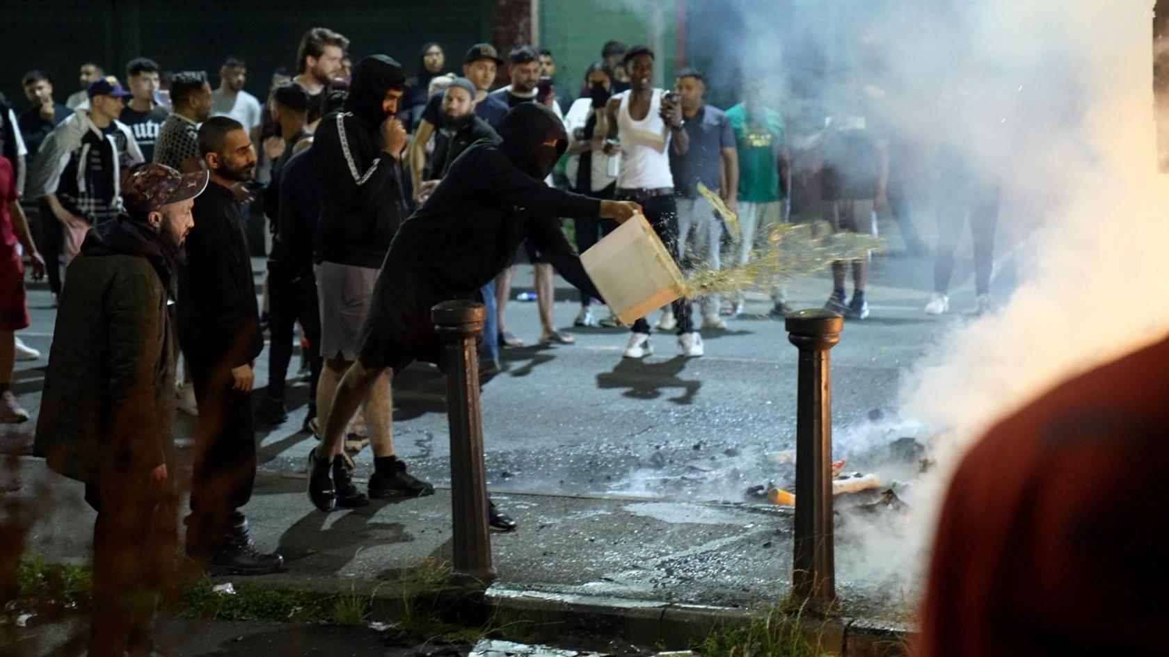 A man in a black hooded top and face mask throws a bucket of water towards something on fire, which is out of the picture. He is surrounded by a crowd of people. 