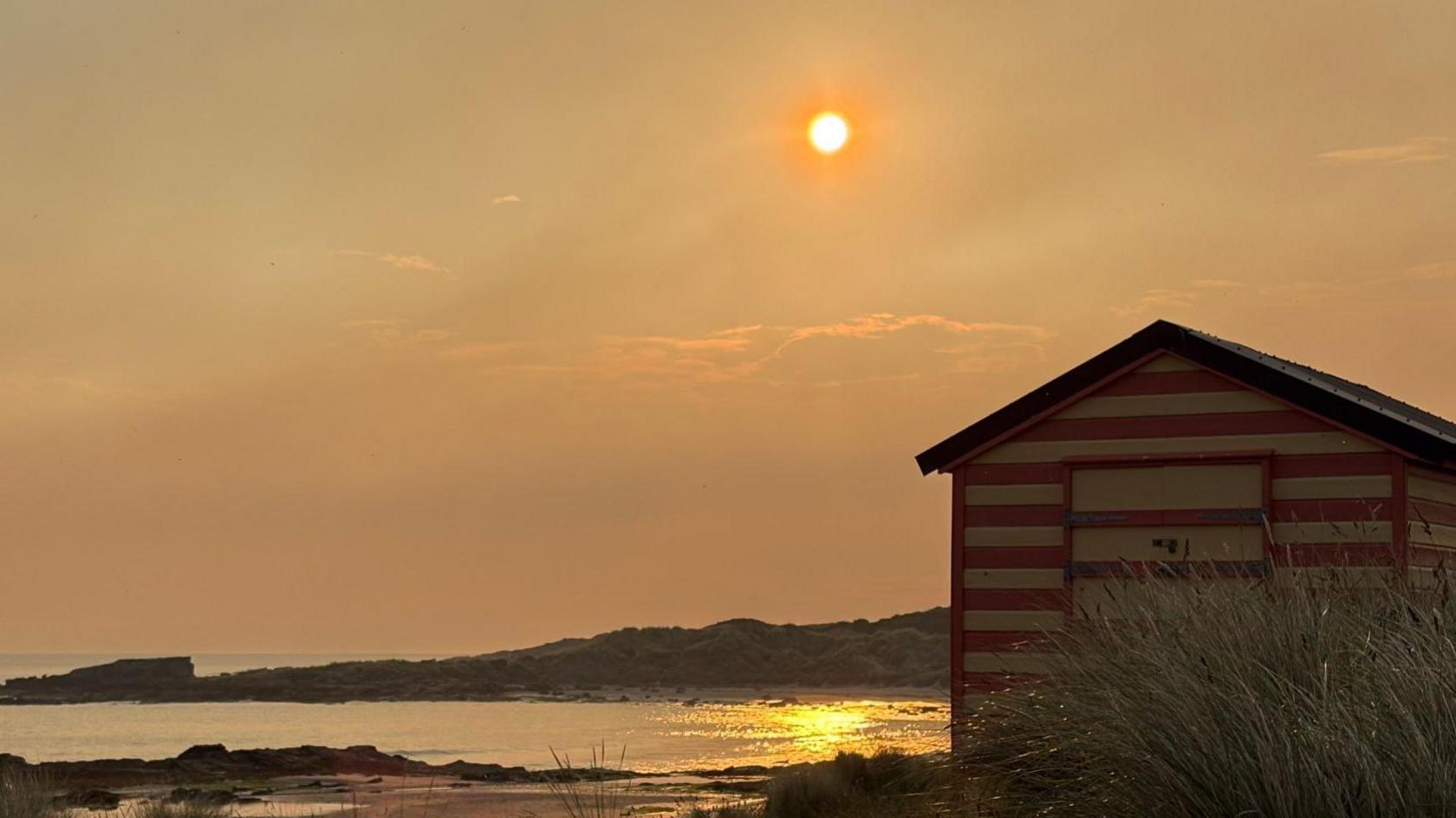 The sun is a bright orange disk in the smoky sky. The colours are reflected in the sea below. There is a beach hut in the foreground. It has a red and white stripy exterior.