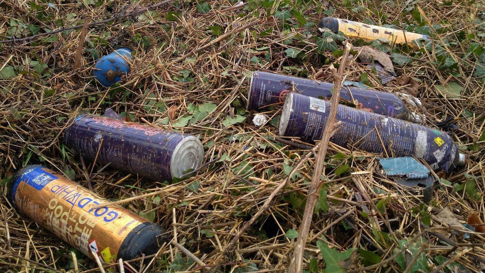 Five blue and gold labelled nitrous oxide canisters discarded in a field beside a road.