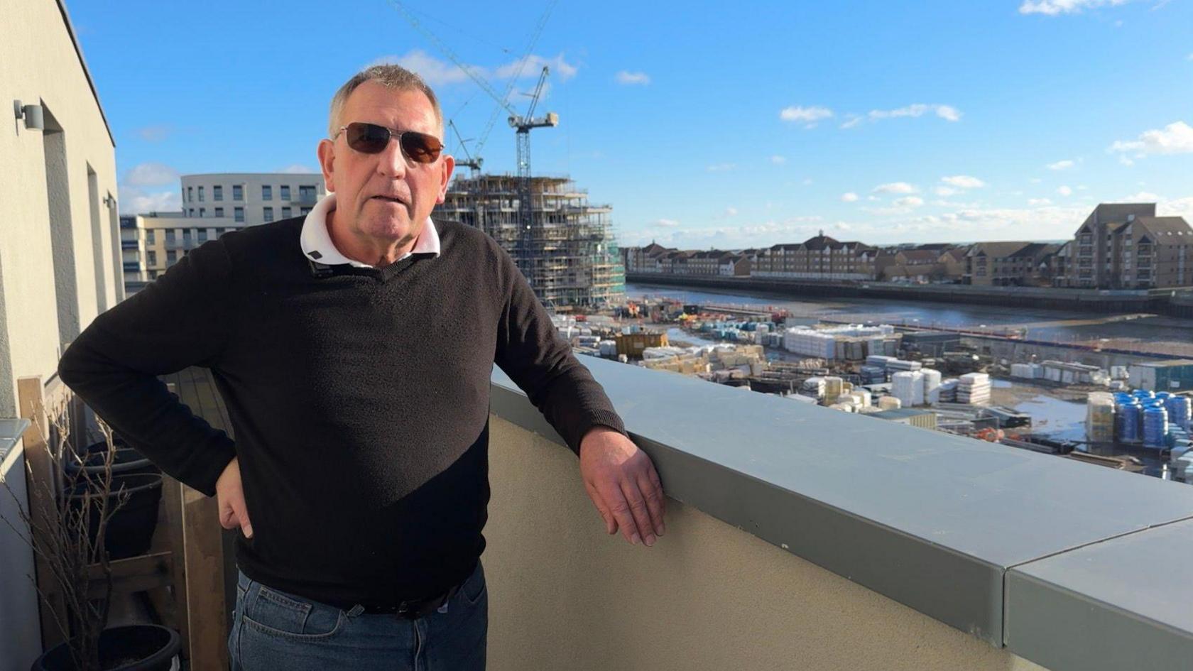 A man with short-cropped grey hair in a black jumper and denim jeans poses on a balcony overlooking an urban river.  