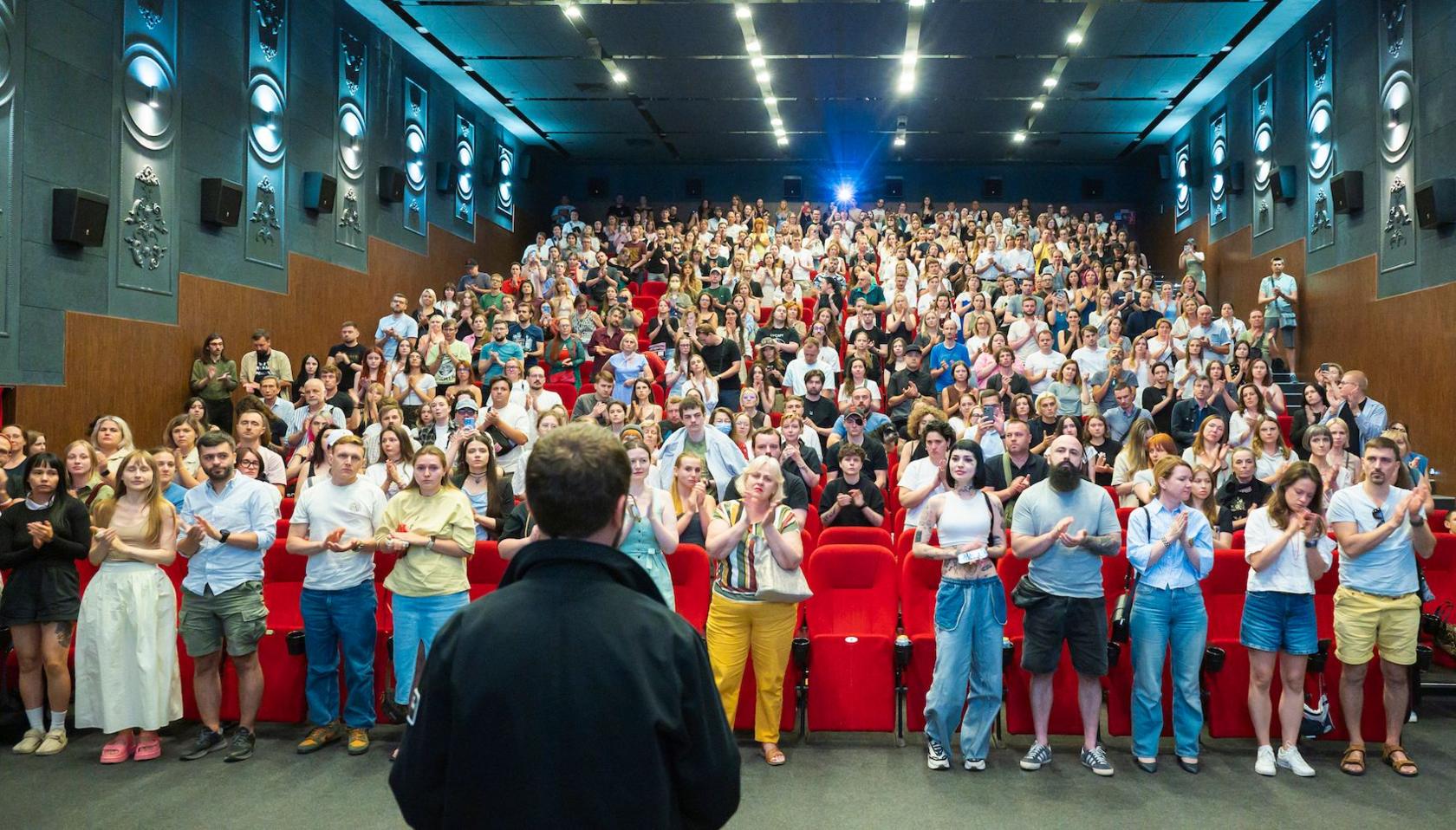 A crowd of people at a cinema in Ukraine are all standing and clapping at the Soundtrack: Ukraine Film Festival on August 31, 2025 . Mstyslav Chernov, the director of the film, is facing away from the camera to the applauding audience. 