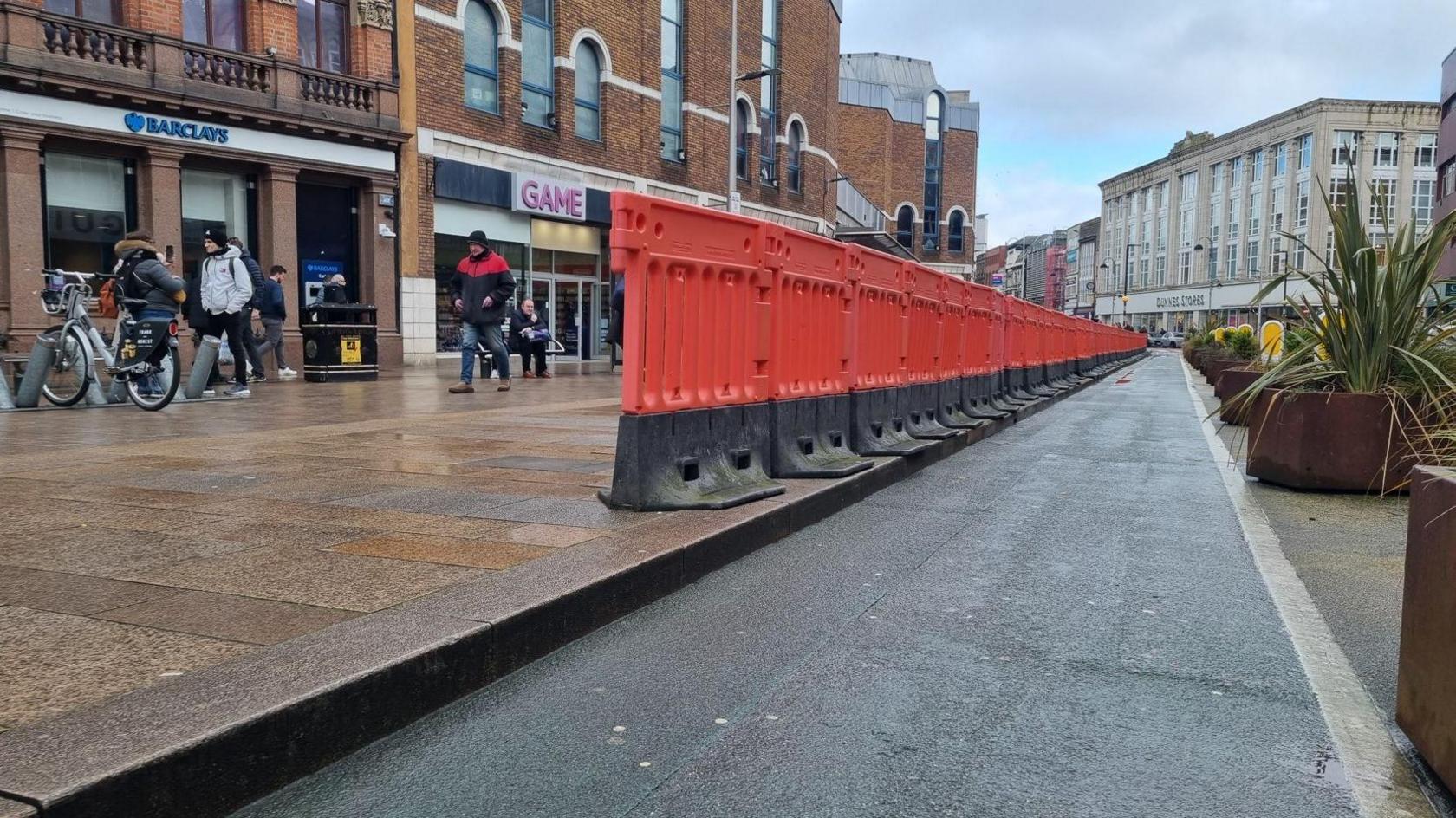 A cycle lane which in lined with an orange barrier on the kerb. People can be seen walking on the footpath past some shops. 