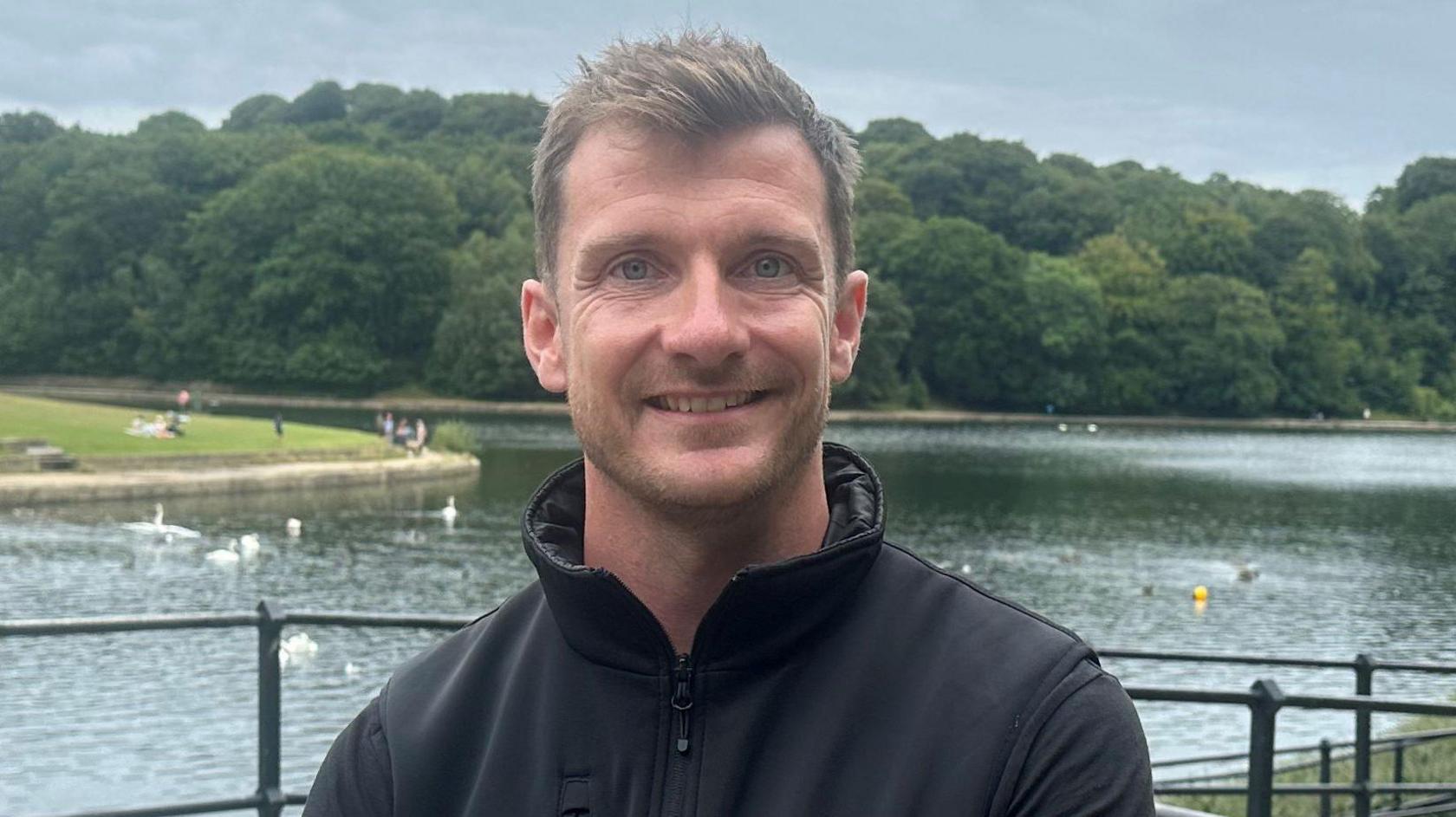 Lewis Peacock, who has short fair hair and is wearing a black fleece. He is smiling in the photo, with a lake in the background.