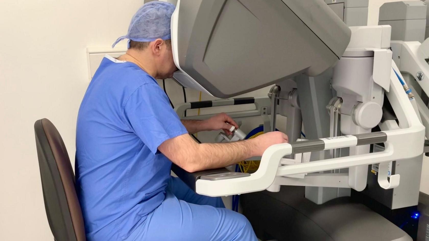 Dr Kevin Burton sits at a console which controls the robot. His head is inside the top of the console and his his eyes are in the viewfinder. We cannot see his face. His hands are operating the levers of the device.
