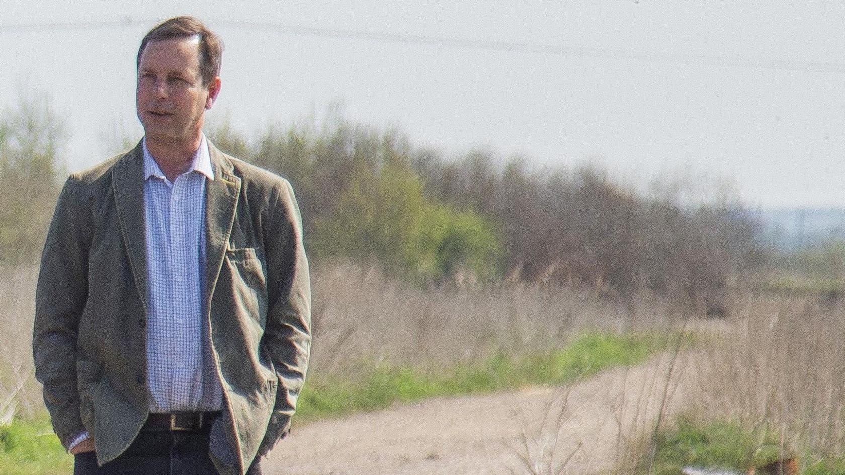 Matt Jackson, standing in a nature reserve, wearing jeans, with a belt, a checked shirt, and jacket, looking slightly away to the left.