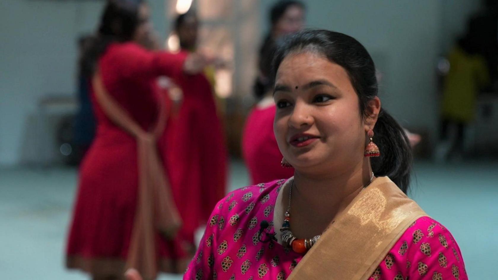 Woman in pink patterned traditional Indian dress with blurred figures dancing in the background