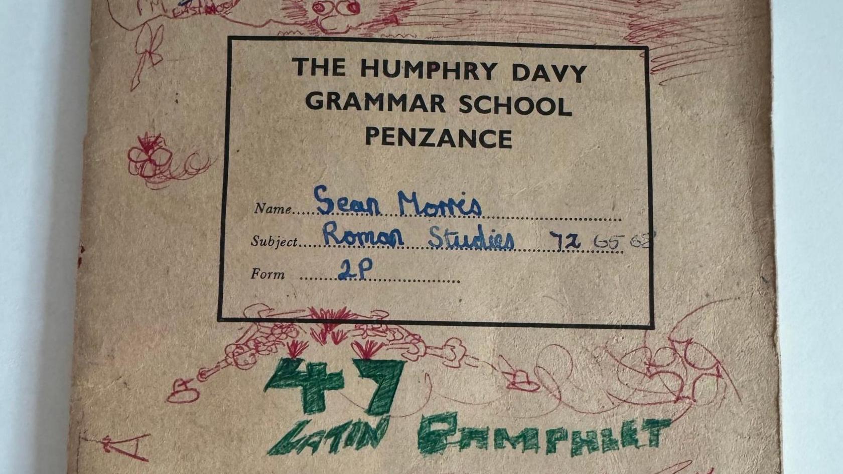 The front cover of the exercise book. It is headed 'The Humphry Davy Grammar School Penzance' and has 'Sean Morris, Roman Studies, 2P' handwritten on it. The rest of the cover has doodles in red and green pen. 