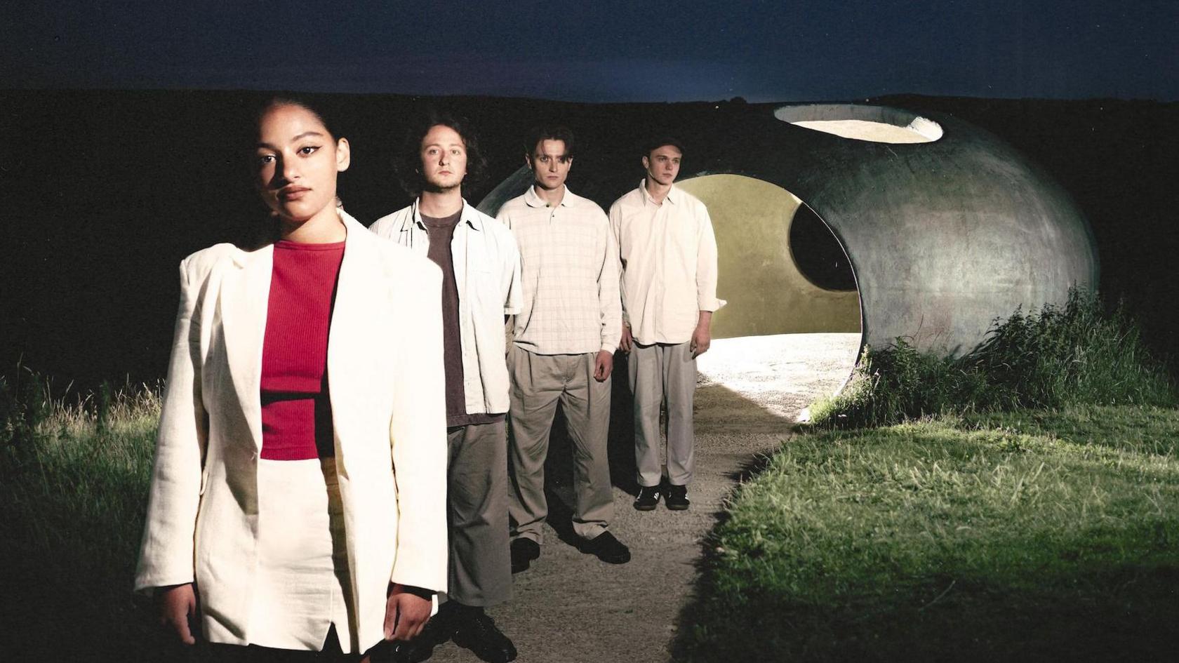 Indie band English Teacher stand in a line while posing for a promotional photo near a hollow stone sculpture, lit from behind