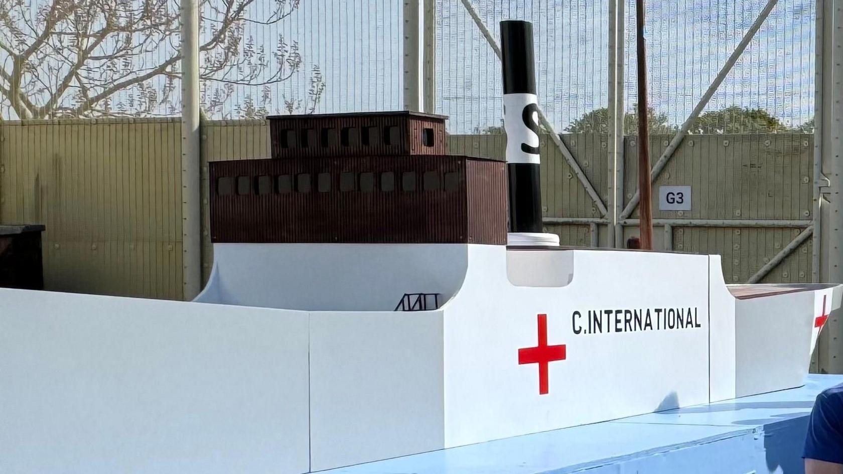 A model of a boat, the SS Vega. It is on a light blue board. The ship is white, with the words C.International on it and a red cross.