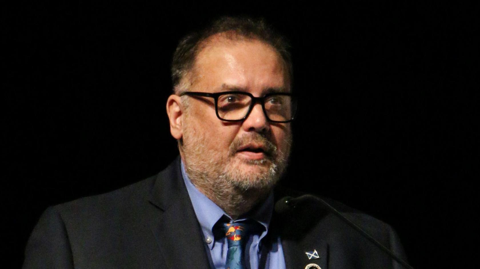A man with a greying beard and black receding hair. He is wearing a blue jacket, shirt and tie