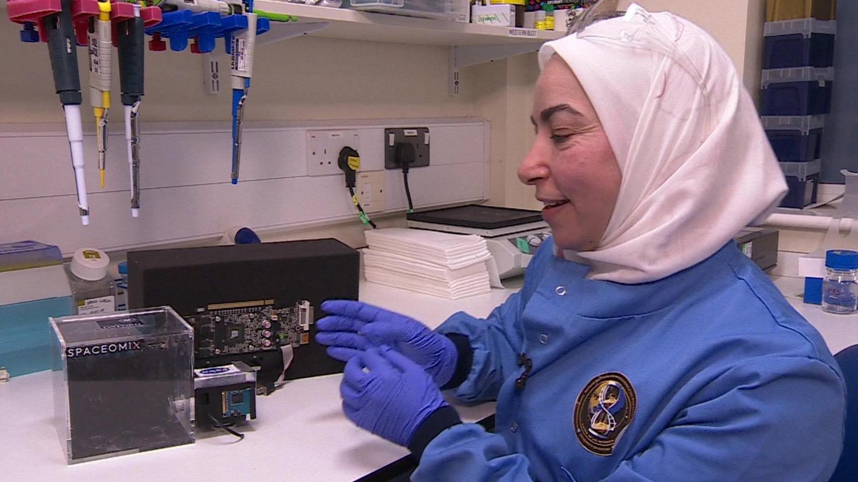 A scientist in a blue jump suit, blue gloves and white head scarf is sitting at a work bench and indicating a piece of technology, which looks like a small plastic box, with her hands 