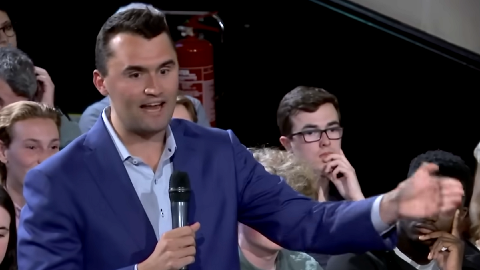 Charlie Kirk in a blue suit and shirt, holding a microphone, with short dark brown hair, gesticulating and talking into a microphone. There are people sitting behind him spectating.