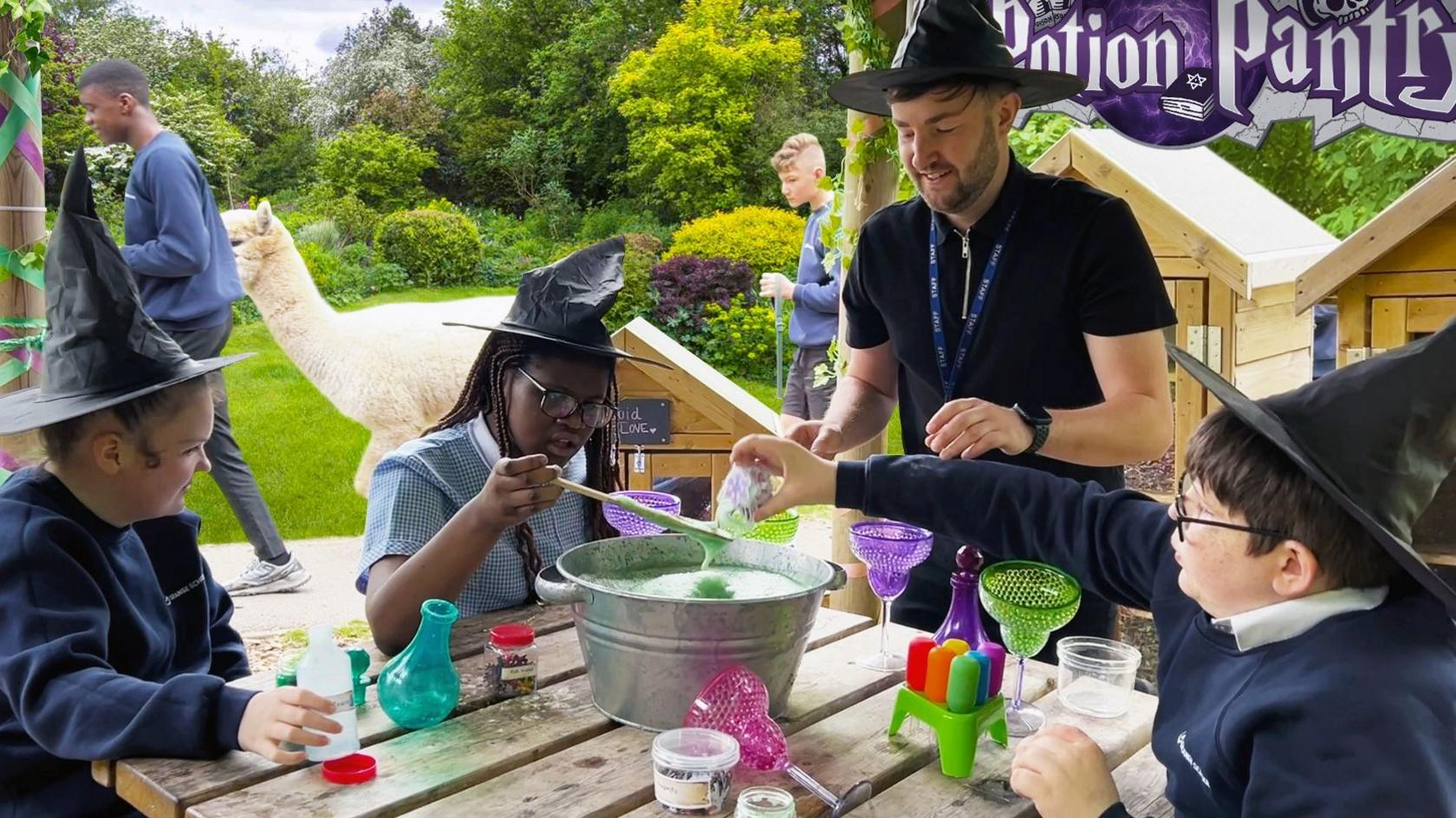 A group of children in witches hats mix colourful liquids in a pot in the middle of the table