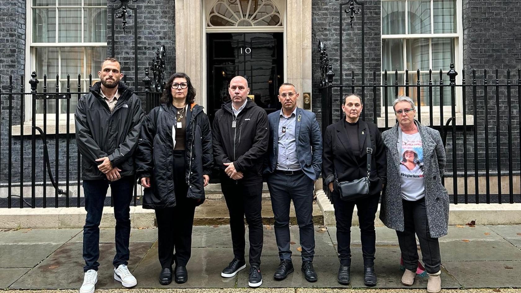 The families of the hostages standing outside the door of Downing Street - there are six people standing in a row with the famous black door behind them. Steve Brisley, Ayelet Svatitzky, and Sharone Lifschitz are all present, with a separate woman wearing a t-shirt with a picture of what is presumably her relative.