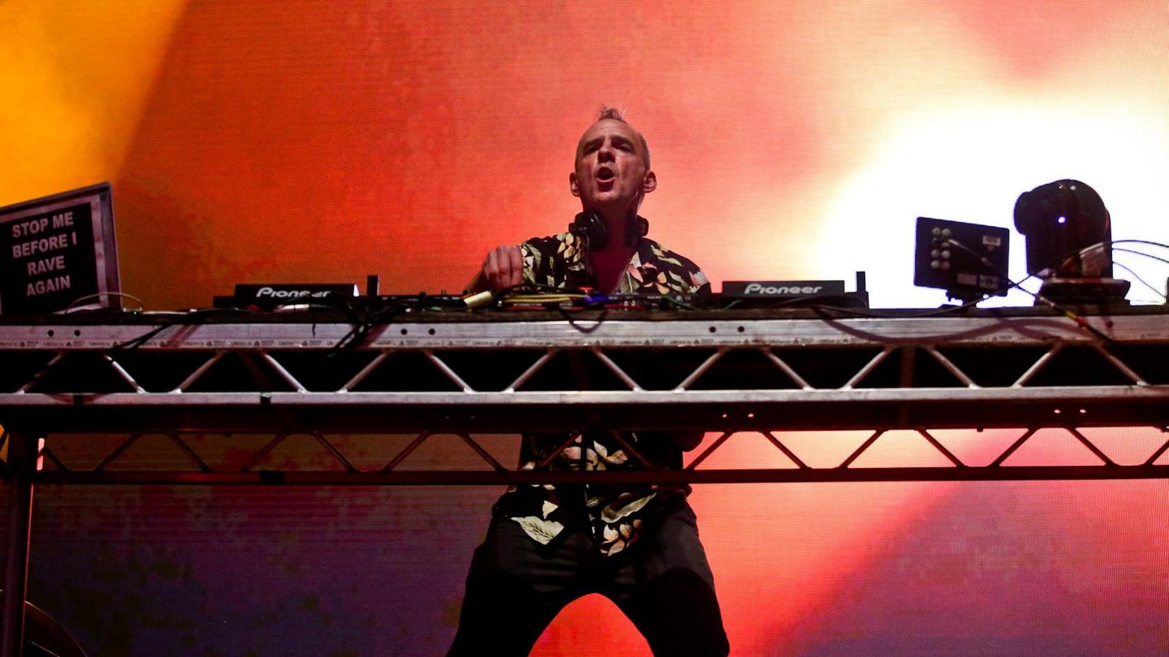 A man in a flowery short sleeved shirt stands behind a set of decks. Behind him is a pink and red screen with flashing lights.