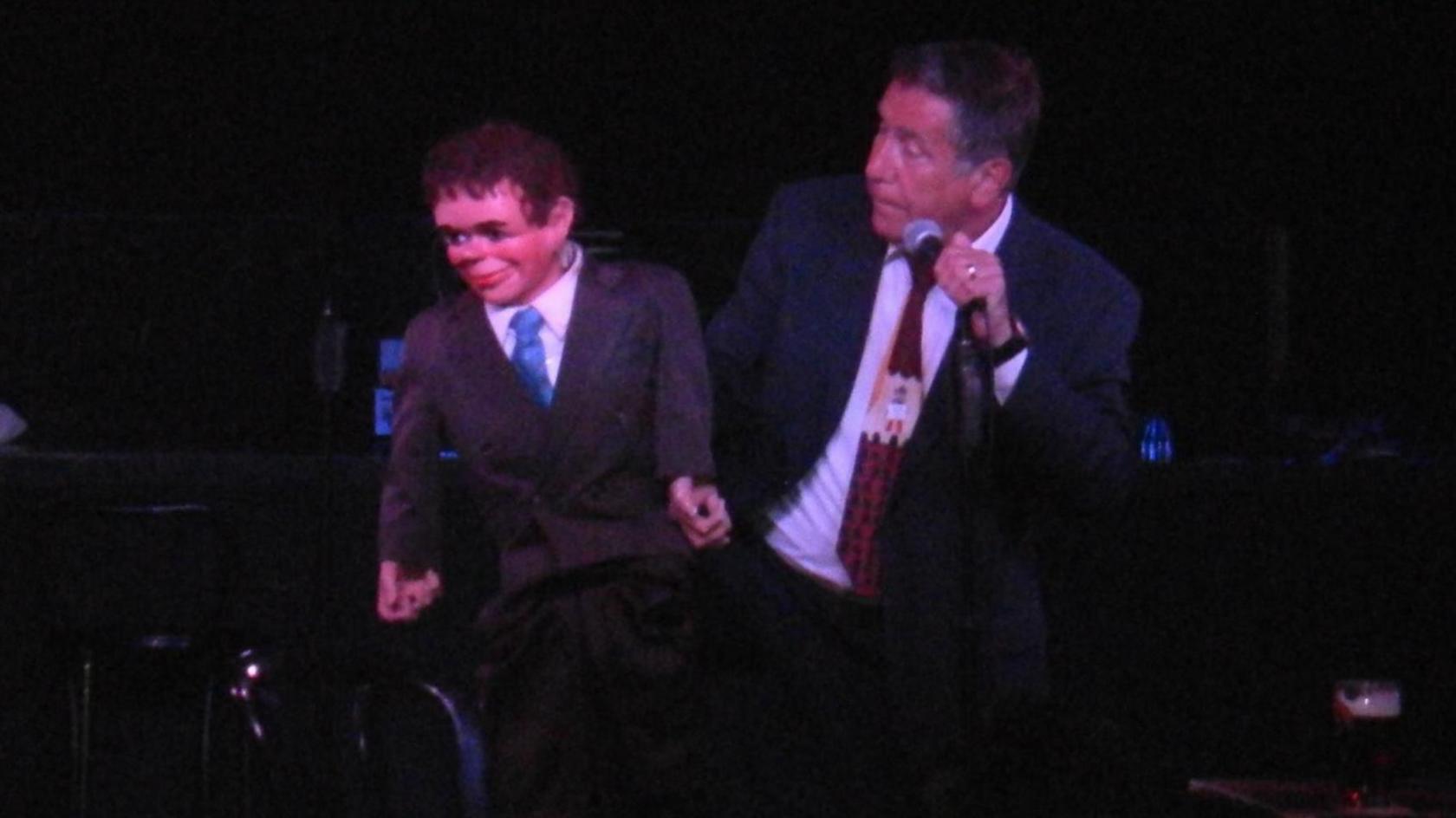 Mr Dennett is holding a microphone while sitting on a chair on stage looking at his ventriloquist's dummy, Chic. Mr Dennett is wearing a dark blue suit with a white shirt and a red, cream and black patterned tie. Chic is wearing a double breasted suit, white shirt and blue tie and is looking away towards the audience. 