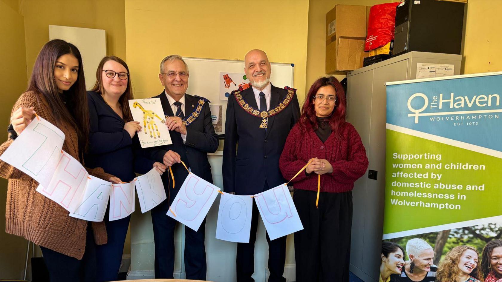 A group of men and women stand holding a paper Thank You sign in front of a white board. A board with The Haven Wolverhampton branding has been placed on the right side of the room.