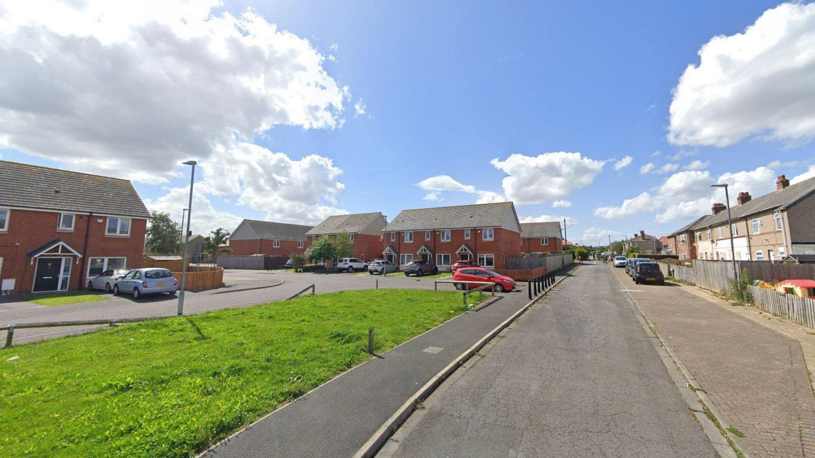 Raby Square looking onto Chatham Gardens. It is a set of interconnected residential streets with lined with red-bricked semi detached houses. There is a green patch of grass in the foreground.