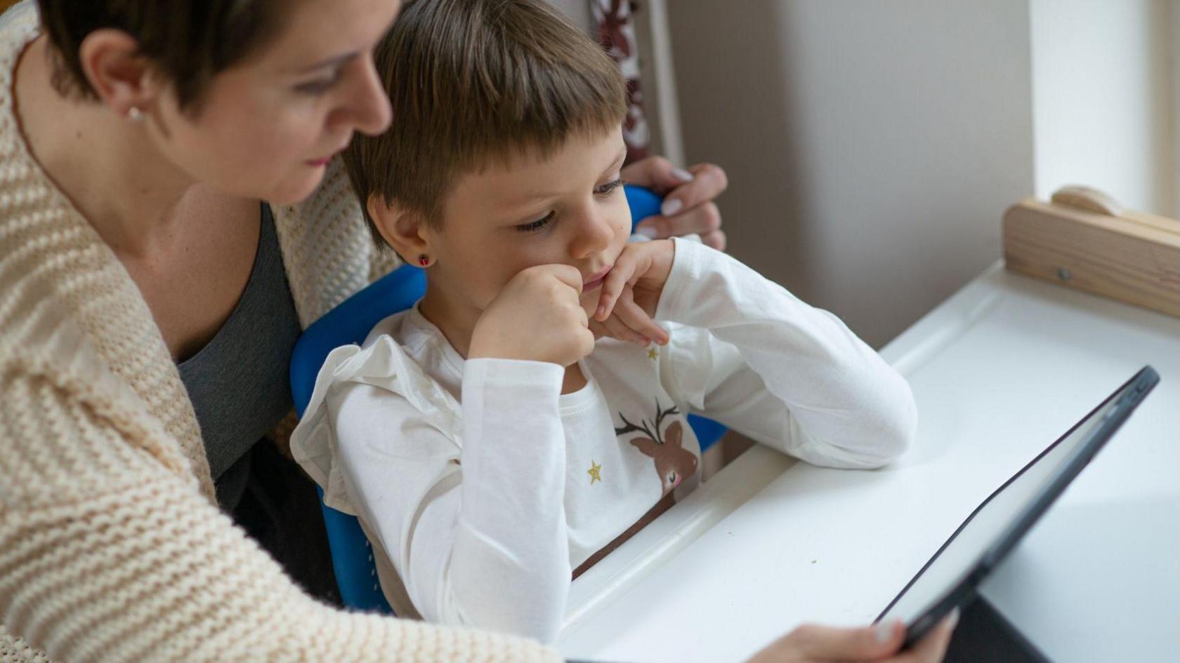 A teacher supporting a young child while they look at a tablet screen together