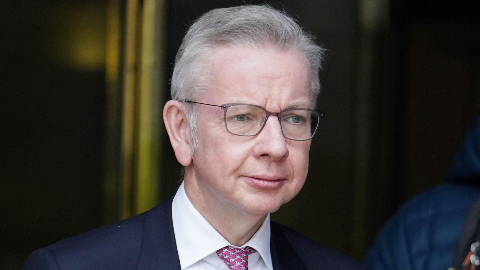 A photo of Michael Gove who was Levelling Up Minister in 2024. He is wearing a suit and tie. He has grey hair and is wearing glasses and is looking to one side of the camera.