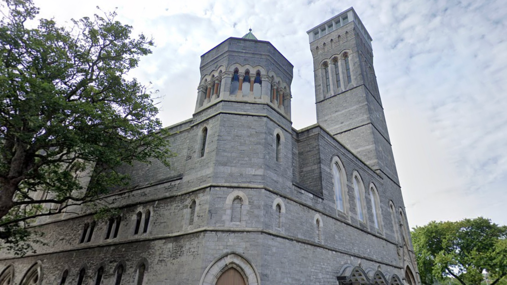 A Google maps image of the Plymouth Guildhall which is a large historic stone building. Trees seen in front on the left and behind on the right, with clouded skies behind.